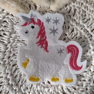 Könnte beinhalten: Ein weißer Einhorn-Flicken aus Filz mit rosa Mähne und Schweif, silbernem Horn und goldenen Hufen. Das Einhorn hat silberne Sternakzente und ist vor einem strukturierten weißen Hintergrund platziert.