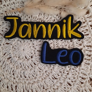 Könnte beinhalten: Zwei gestickte Patches mit Namen. Der erste Patch ist schwarz mit gelben gestickten Buchstaben, die "Jannik" buchstabieren. Der zweite Patch ist schwarz mit blauen gestickten Buchstaben, die "Leo" buchstabieren.