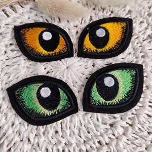Könnte beinhalten: Vier gestickte Augen-Patches. Zwei Patches haben gelbe und orangefarbene Augen, und zwei haben grüne Augen. Jedes Auge hat eine schwarze Umrandung und eine weiße Pupille. Die Patches liegen auf einer weißen, gewebten Oberfläche.