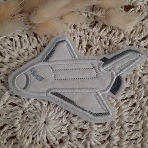 Könnte beinhalten: Weißer Stoff-Space-Shuttle-Patch mit grauen Stickdetails. Der Patch ist so gestaltet, dass er wie ein Space Shuttle mit einer spitzen Nase und Flügeln aussieht.