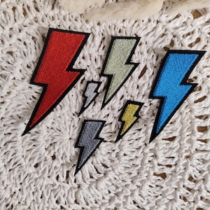Könnte beinhalten: Eine Sammlung von gestickten Blitz-Patches in verschiedenen Farben, darunter Rot, Blau, Gelb und Grau. Jeder Patch hat einen schwarzen Rand. Die Patches sind auf einer weißen, strukturierten Oberfläche angeordnet.