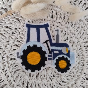 Könnte beinhalten: Ein gestickter Traktor-Patch in Blau und Weiß mit gelben Rädern. Der Patch ist perfekt, um Ihren Projekten einen Hauch von Bauernhof-Charme zu verleihen.