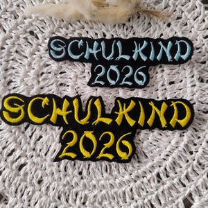 Könnte beinhalten: Zwei schwarze Stoffaufnäher mit gesticktem Text. Der obere Aufnäher zeigt hellblaue Schrift mit dem Text SCHULKIND 2026. Der untere Aufnäher zeigt gelbe Schrift mit dem Text SCHULKIND 2026. Beide Aufnäher haben eine wellige, unregelmäßige Form.