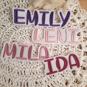 Könnte beinhalten: Vier Filzpatches mit Namen, die in verschiedenen Farben gestickt sind. Die Namen sind "Emily" in Lila, "Leni" in Rosa, "Mila" in Rosa und "Ida" in Rot.