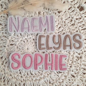 Könnte beinhalten: Drei gestickte Stoff-Patches mit den Namen "Naemi", "Elyas" und "Sophie" in rosa, braun und rosa Garn. Die Patches befinden sich auf einer weißen Häkel-Oberfläche.