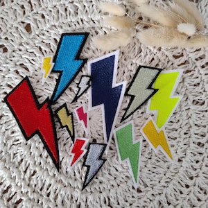 Könnte beinhalten: Eine Sammlung von gestickten Blitz-Aufnähern in verschiedenen Größen und Farben, darunter Rot, Blau, Marineblau, Gelb, Grün, Silber und Pink. Jeder Aufnäher hat einen markanten schwarzen Rand und eine weiße Stoffkante mit detaillierten Stichen.