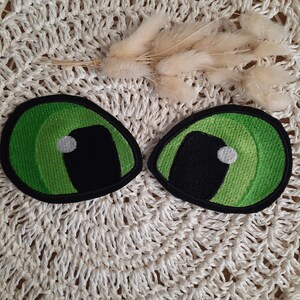 Könnte beinhalten: Zwei grüne und schwarze gestickte Filzaugen mit weißen Pupillen. Die Augen befinden sich auf einer weißen gehäkelten Oberfläche.