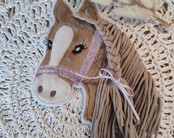 以下が含まれることがあります： 茶色、ベージュ、白の色合いのぬいぐるみ馬の頭で、ピンクのグリッターのハルターが付いています。馬は三つ編みのたてがみと、端に白いリボンが付いています。馬は白いかぎ針編みの表面にあります。