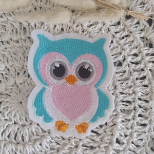 Könnte beinhalten: Gestickter Eulenaufnäher in Blau, Rosa und Weiß. Die Eule hat große Augen und einen orangefarbenen Schnabel und Füße. Der Aufnäher ist für Kleidung oder Accessoires gedacht.