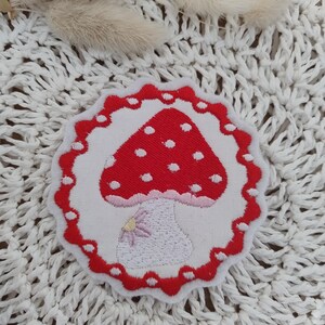 Könnte beinhalten: Roter und weißer gestickter Pilz-Patch mit weißen Punkten. Der Pilz ist von einem roten, gewellten Rand mit weißen Punkten umgeben.