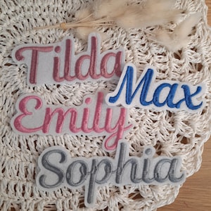 Könnte beinhalten: Vier gestickte Stoffpatches mit den Namen: Tilda, Emily, Max und Sophia. Die Patches sind weiß mit rosa, blauen und grauen Stickereien.