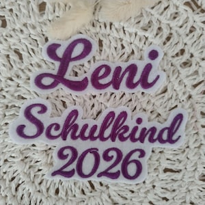 Könnte beinhalten: Zwei weiße Stoffaufnäher mit violetter Stickerei. Der obere Aufnäher zeigt den Namen Leni in Schreibschrift. Der untere Aufnäher zeigt den Text Schulkind und die Jahreszahl 2026 im gleichen violetten Schreibschrift-Stickstil.