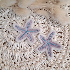 Könnte beinhalten: Zwei gestickte Seestern-Patches. Die Seesterne sind lavendelfarben mit hellblauen Akzenten und weißen Rändern. Die Patches befinden sich auf einem weißen, gewebten Hintergrund mit getrockneten beigen Pflanzen im Hintergrund.