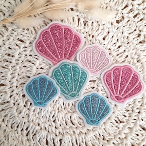 Könnte beinhalten: Fünf glitzernde Muschel-Patches in verschiedenen Schattierungen von Rosa, Blau und Türkis. Die Patches sind auf eine weiße gehäkelte Oberfläche genäht.