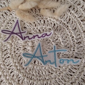 Könnte beinhalten: Zwei gestickte Namensaufnäher. Einer ist lila und trägt den Namen "Anna". Der andere ist hellblau und trägt den Namen "Anton". Beide Aufnäher befinden sich auf einem weißen Hintergrund mit einem strukturierten Muster.