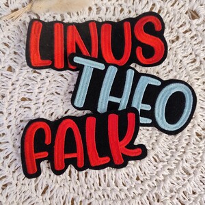 Könnte beinhalten: Drei gestickte Aufnäher mit den Namen "LINUS" in Rot, "THEO" in Hellblau und "FALK" in Rot. Jeder Name befindet sich auf einem schwarzen Hintergrund mit weißem Rand. Die Aufnäher werden auf einer weißen, strukturierten Oberfläche präsentiert.