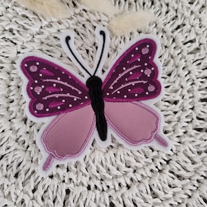 Könnte beinhalten: Eine rosa und violette Schmetterlingsapplikation mit schwarzen Akzenten und weißen Nähten. Der Schmetterling befindet sich auf einem weißen Häkelhintergrund.