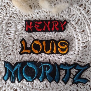 Könnte beinhalten: Drei gestickte Patches mit Namen in einer stilisierten Schrift. Der obere Patch ist rot und sagt "HENRY". Der mittlere Patch ist gelb und sagt "LOUIS". Der untere Patch ist blau und sagt "MORITZ".
