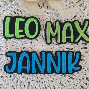 Könnte beinhalten: Drei gestickte Stoff-Patches mit den Namen: Leo, Max und Jannik. Die Patches sind schwarz mit grüner und blauer Schrift.