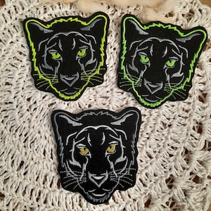 Könnte beinhalten: Drei schwarze Pantherkopf-Patches mit neongrünen Umrissen. Die Patches sind gestickt und haben ein detailliertes Design.
