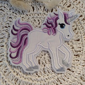 Könnte beinhalten: Weißer Einhorn-Applikator mit einer lila und pinkfarbenen Mähne und einem Schwanz. Das Einhorn hat ein silbernes Horn und Hufe.