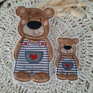 Könnte beinhalten: Zwei braune Teddybär-Applikationen mit blau-weiß gestreiften Latzhosen und roten Herzen. Der größere Bär steht, der kleinere sitzt.