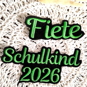 Könnte beinhalten: Zwei gestickte Aufnäher mit grünem Text auf schwarzem Hintergrund. Der obere Aufnäher lautet "Fiete". Der untere Aufnäher lautet "Schulkind 2026". Die Aufnäher befinden sich auf einer weißen, strukturierten Oberfläche.