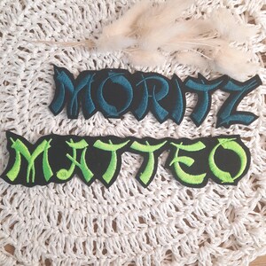 Könnte beinhalten: Zwei gestickte Patches mit den Namen "MORITZ" und "MATTEO" in schwarzer, grüner und blauer Schrift. Die Patches befinden sich auf einer weißen Häkelunterlage.