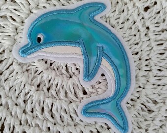 Könnte beinhalten: Ein blauer, schimmernder Delfin-Patch mit weißen Nähten. Der Patch befindet sich auf einer weißen gehäkelten Oberfläche.