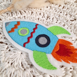 Könnte beinhalten: Ein gestickter Raketen-Patch in Blau und Grün mit orangefarbenen und roten Flammen. Die Rakete hat einen schwarzen und grauen Kreis in der Mitte.