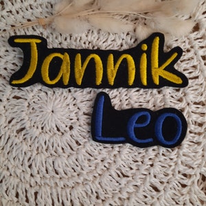 Könnte beinhalten: Zwei gestickte Patches mit Namen auf einer weißen gehäkelten Oberfläche. Der obere Patch ist schwarz mit gelben gestickten Buchstaben, die "Jannik" buchstabieren. Der untere Patch ist schwarz mit blauen gestickten Buchstaben, die "Leo" buchstabieren.