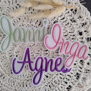 Könnte beinhalten: Gestickte Namensaufnäher in Grün, Rosa und Lila. Die Namen "Janni", "Inga" und "Agnes" sind in einer Schreibschrift mit weißem Hintergrund dargestellt. Die Aufnäher liegen auf einer weißen, strukturierten Oberfläche mit getrockneten Blumen.