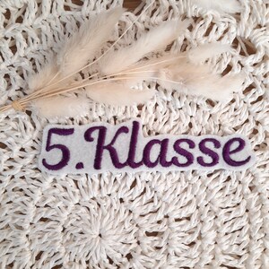 Könnte beinhalten: Weißer Filz-Patch mit violett gesticktem Text "5. Klasse".