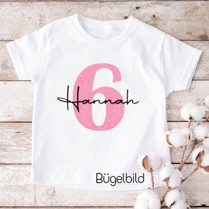 Könnte beinhalten: Weißes T-Shirt mit einer pinkfarbenen Zahl 6 und dem Namen "Hannah" in schwarzer Schrift. Der Text "Bügelbild" ist unter dem Design gedruckt.