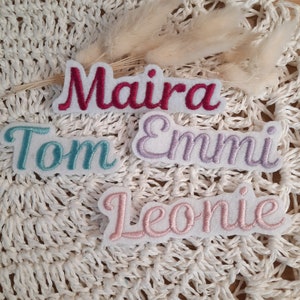 Könnte beinhalten: Vier gestickte Stoff-Patches mit Namen darauf. Die Namen sind Maira, Tom, Emmi und Leonie. Die Patches befinden sich auf einer weißen gehäkelten Oberfläche.