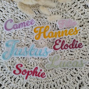 Könnte beinhalten: Ein Set aus sieben bunten, gestickten Bügeletiketten mit Namen in Schreibschrift. Die Namen sind: Camee, Fillin, Hannes, Justus, Elodie, Lucas und Sophie.