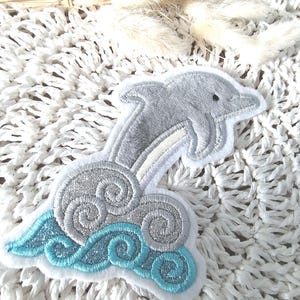 Könnte beinhalten: Ein gestickter grauer Delfin springt aus blauen und silbernen Wellen. Der Delfin ist weiß umrandet und hat ein schwarzes Auge. Die Wellen sind glitzernd.