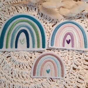 Könnte beinhalten: Drei gestickte Regenbogen-Patches mit einem Herz in der Mitte jedes. Die Regenbögen sind in Blautönen, Grüntönen und Rosatönen gehalten. Die Herzen sind weiß, blau und lila.