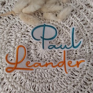 Könnte beinhalten: Zwei gestickte Namensaufnäher, "Paul" in Türkis und "Leander" in Orange, auf weißem Filz. Die Aufnäher sind auf einer strukturierten weißen Oberfläche platziert, mit Trockenblumen im Hintergrund. Die Schrift ist kursiv.