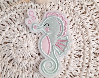 May include: A light blue and pink embroidered seahorse with a whimsical expression, blowing bubbles. The seahorse is surrounded by a white crocheted background.