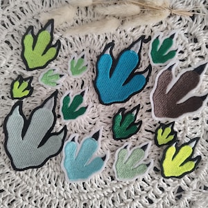 Könnte beinhalten: Eine Sammlung gestickter Dinosaurier-Fußabdruck-Patches in verschiedenen Größen und Farben. Die Patches weisen Grüntöne, Blau- und Brauntöne mit schwarzen Krallen und weißen Rändern auf. Die Patches sind auf einer weißen, strukturierten Oberfläche angeordnet.