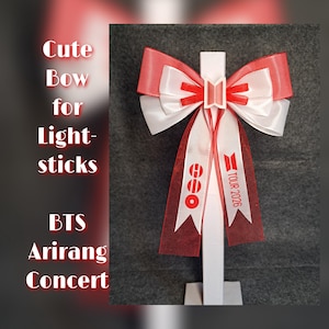 Può includere: Un fiocco rosso e bianco per light stick, con la scritta "TOUR 2026" e "BTS Arirang Concert". Il fiocco è fatto di nastro e ha un logo al centro.