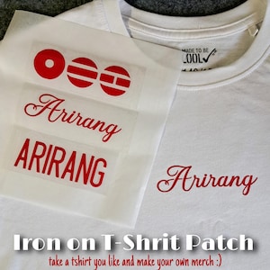 Könnte beinhalten: Ein weißes T-Shirt mit einem roten Aufbügel-Transfer mit dem Wort Arirang in Schreibschrift und Blockbuchstaben sowie drei roten kreisförmigen Symbolen. Ein passendes rotes Arirang-Logo ist bereits auf der Brust des weißen T-Shirts angebracht.