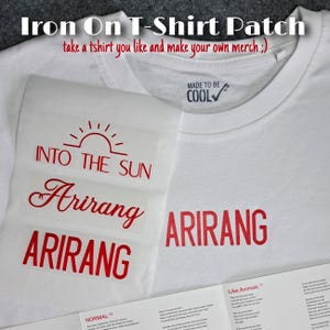 Könnte beinhalten: Ein weißes T-Shirt mit einem roten Bügelbild mit dem Text INTO THE SUN, Arirang und ARIRANG. Das T-Shirt hat ein Nackenetikett mit der Aufschrift MADE TO BE COOL. Ein Bogen mit roten Bügeltransfers mit demselben Text liegt auf dem Stoff.