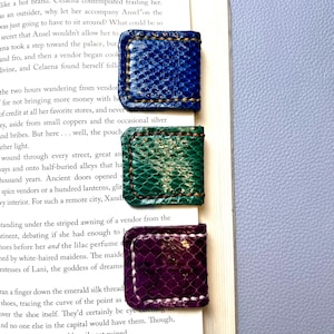 May include: Three square magnetic bookmarks with a textured snakeskin pattern in blue, green, and purple. Each bookmark features visible stitching along the edges and is clipped onto the side of an open book page with printed text.