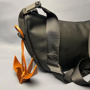 Puede incluir: Bolso bandolera negro con hebilla plateada y un charm de grulla origami de cuero marrón. El bolso tiene una correa ancha negra y un diseño curvo. El charm está hecho de cuero plegado.