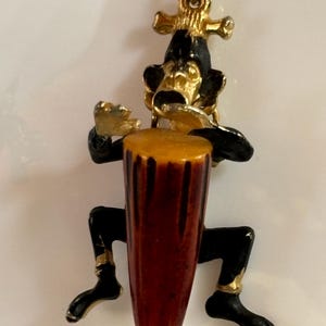 Könnte beinhalten: Eine Halskette mit einem schwarzen und goldenen Anhänger, der einen Affen zeigt, der Trommel spielt. Die Trommel ist aus Bernstein und hat eine braune und gelbe Farbe. Der Affe trägt eine goldene Krone und einen goldenen Ring am Finger.