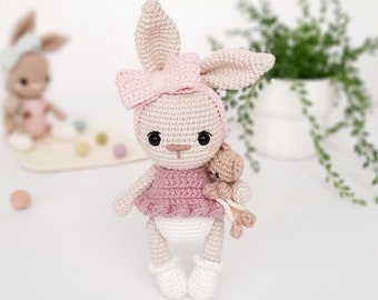 May include: A crocheted rabbit doll with long ears, wearing a pink headband with a bow, a dusty pink dress, and white shoes. The rabbit has black eyes and holds a small, light brown crocheted teddy bear in one arm, featuring detailed yarn stitching throughout.