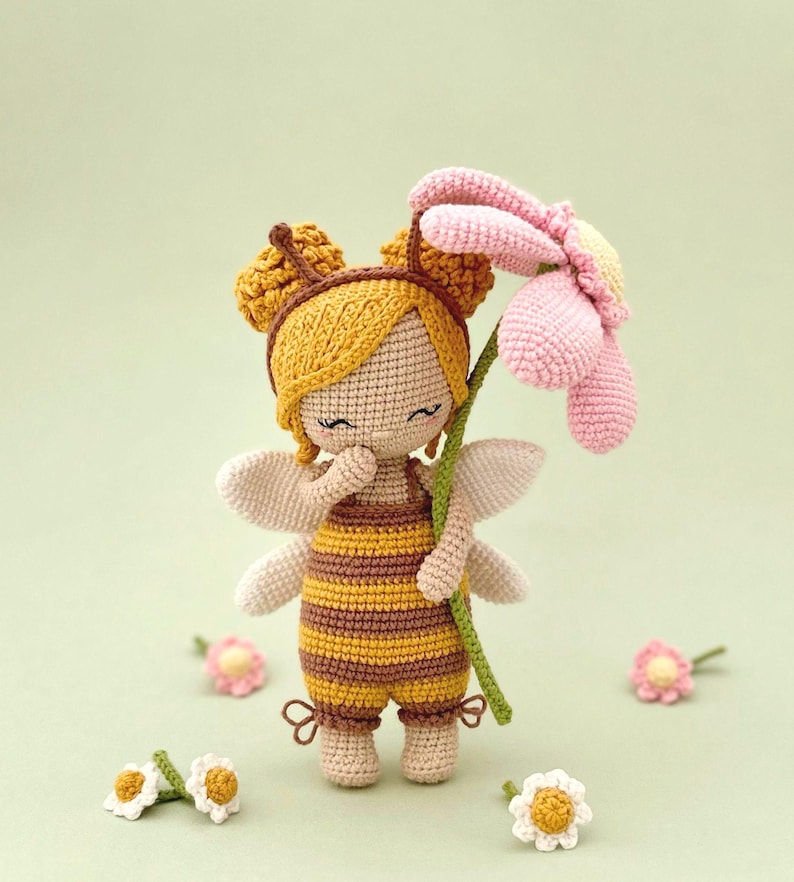 Peut inclure: Une poup&eacute;e crochet&eacute;e v&ecirc;tue d'une tenue d'abeille &agrave; rayures jaunes et brunes avec de petites ailes, tenant une grande fleur rose et jaune. La poup&eacute;e a des cheveux jaunes coiff&eacute;s en deux chignons et est entour&eacute;e de quatre petites fleurs en crochet.