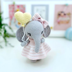 Peut inclure: Un &eacute;l&eacute;phant en crochet gris portant une robe ray&eacute;e rose et blanche et un n&oelig;ud rose. L'&eacute;l&eacute;phant tient un ballon jaune en forme de c&oelig;ur.